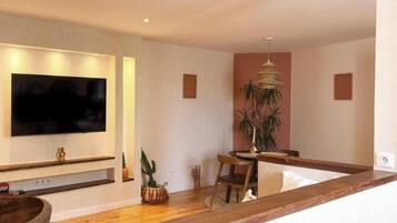 Una sala moderna con televisor de pantalla plana montado en la pared, un comedor con mesa y sillas de madera, y una planta grande en maceta.