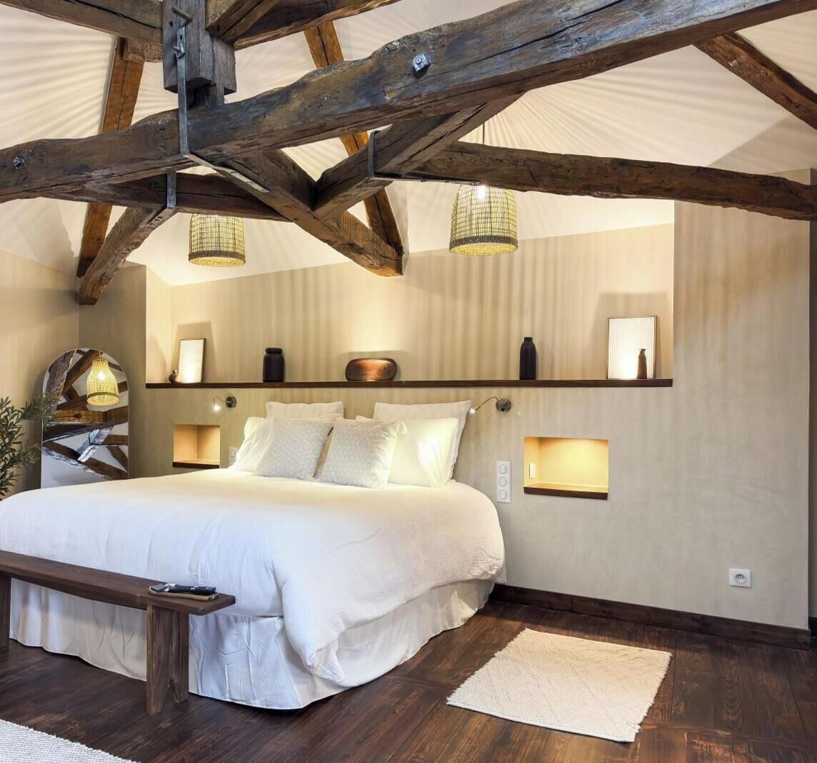 Une chambre avec un grand lit, des poutres apparentes et des luminaires fixés au mur.