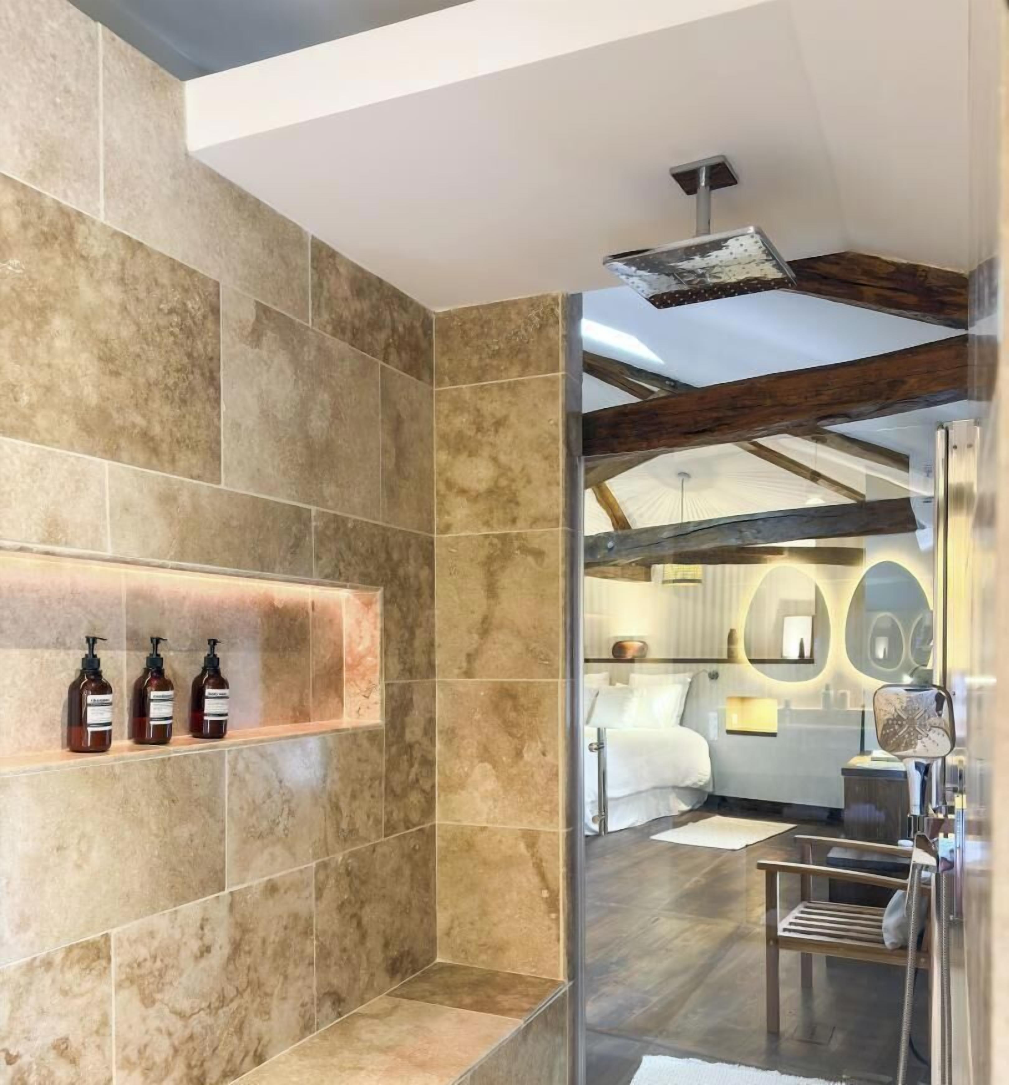 A modern bathroom with a shower, a shelf with toiletries, and a view into a bedroom with a bed and a nightstand.