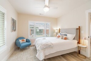 Une chambre à coucher comprenant un lit, une chaise, une fenêtre avec des stores, un ventilateur de plafond et une table de chevet avec une lampe.