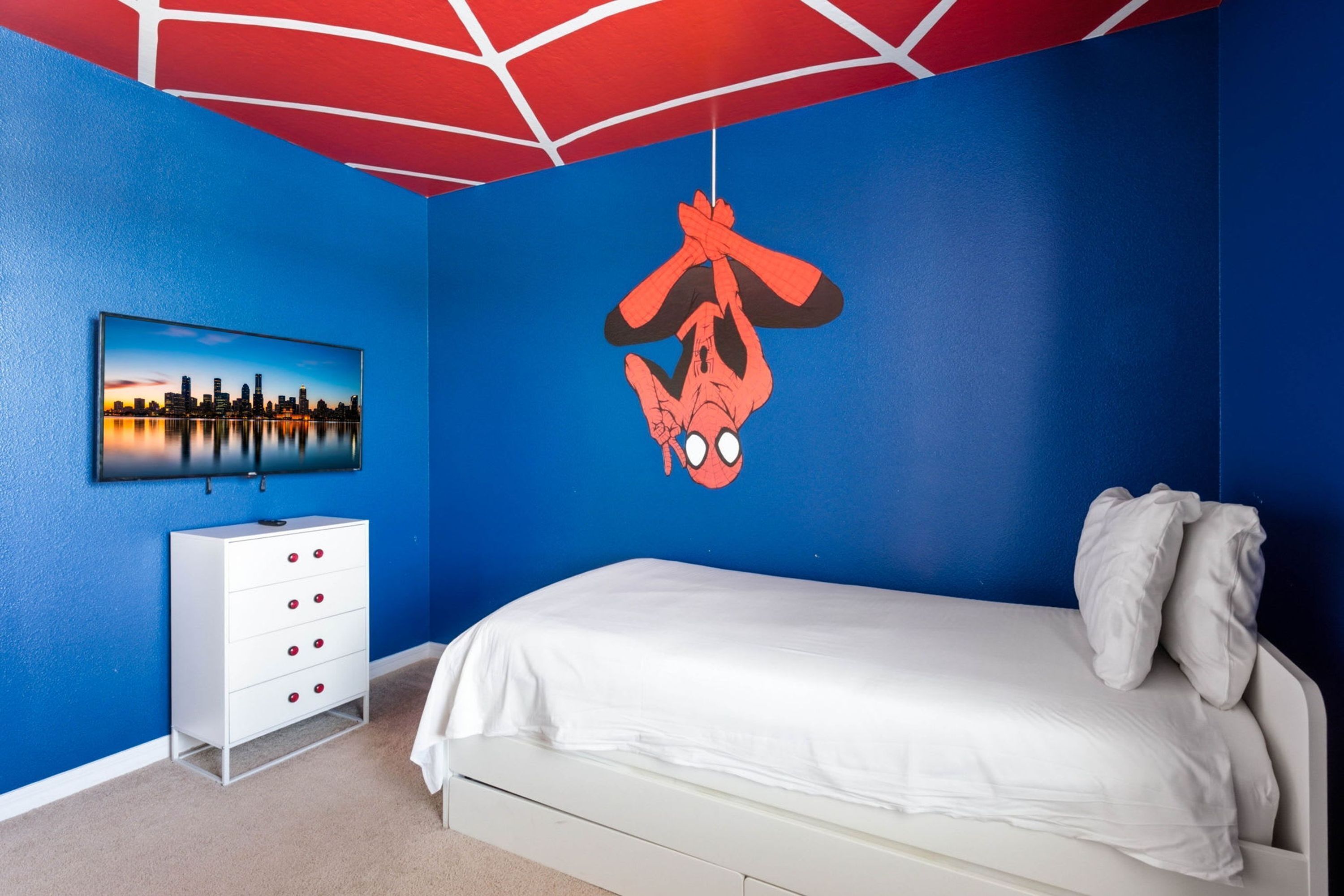 A bedroom with a white bed, a white dresser, a cityscape painting, and a Spider-Man decal on the wall.