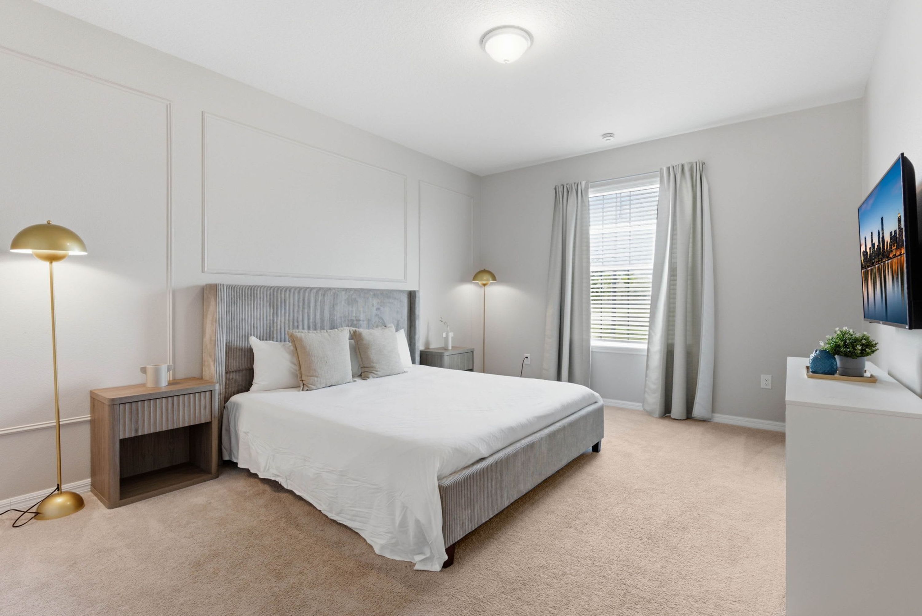 Een moderne slaapkamer met een groot bed, bedlampjes, een nachtkastje, een raam met gordijnen en een stadsgezichtschilderij aan de muur.