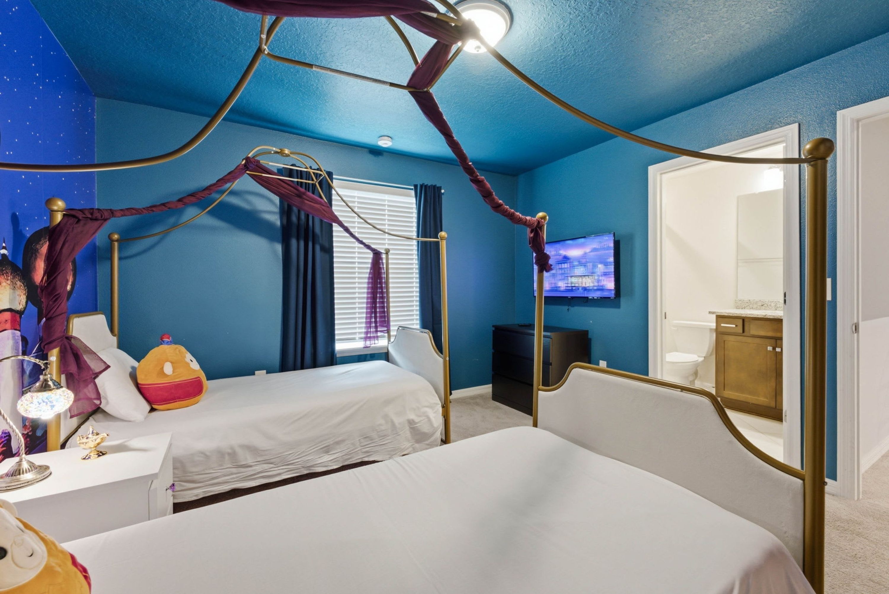 A room with a canopy bed, a television, a desk with a lamp, and a window with curtains.