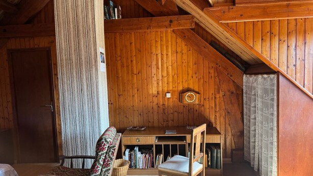 A cozy attic room with wooden walls, a desk, a chair, and a bookshelf.