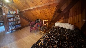 A cozy attic bedroom with a wooden ceiling, a bookshelf, a chair, and a bed with a floral bedspread.