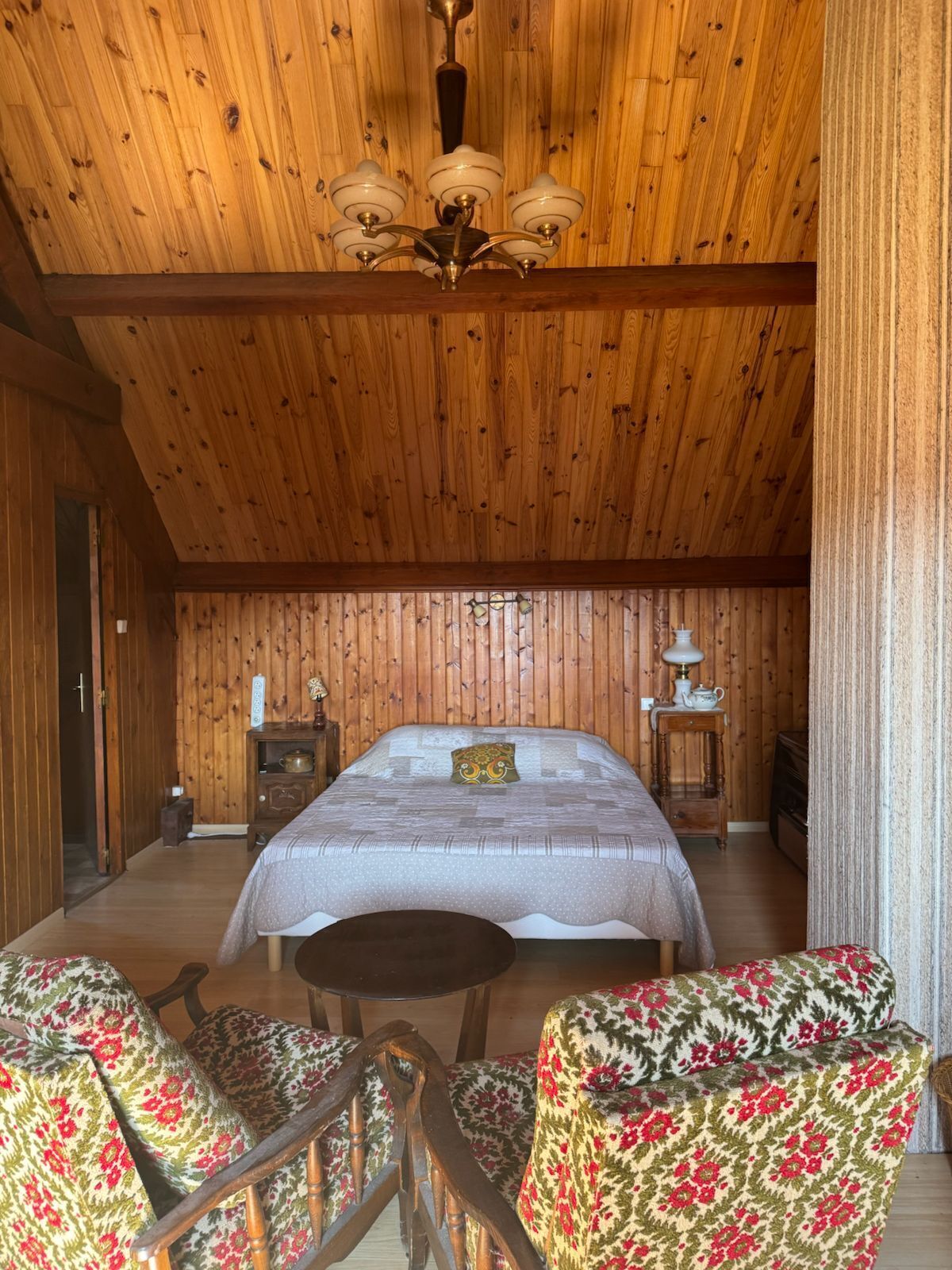 Quarto com painéis de madeira, uma cama, duas cadeiras, uma mesinha e um criado-mudo com luminária.