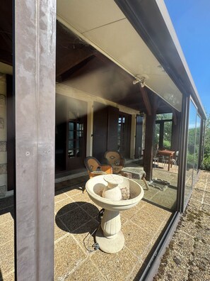 Un patio couvert avec une table et des chaises en pierre, une structure vitrée et une vue sur l’extérieur.