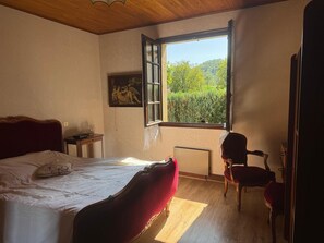 Un dormitori amb un llit gran, una cadira, una taula i una finestra que dona a un espai verd.