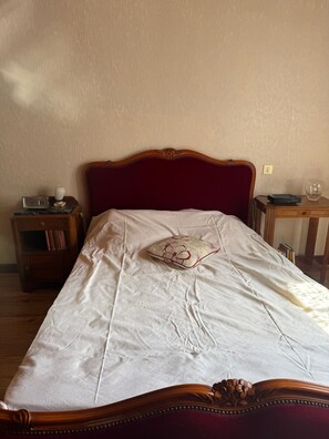 En säng med ett vitt lakan och en prydnadskudde med blommönster, ett träbord vid sängen med en klocka och böcker, och ett litet bord med en lampa och ett glas.
