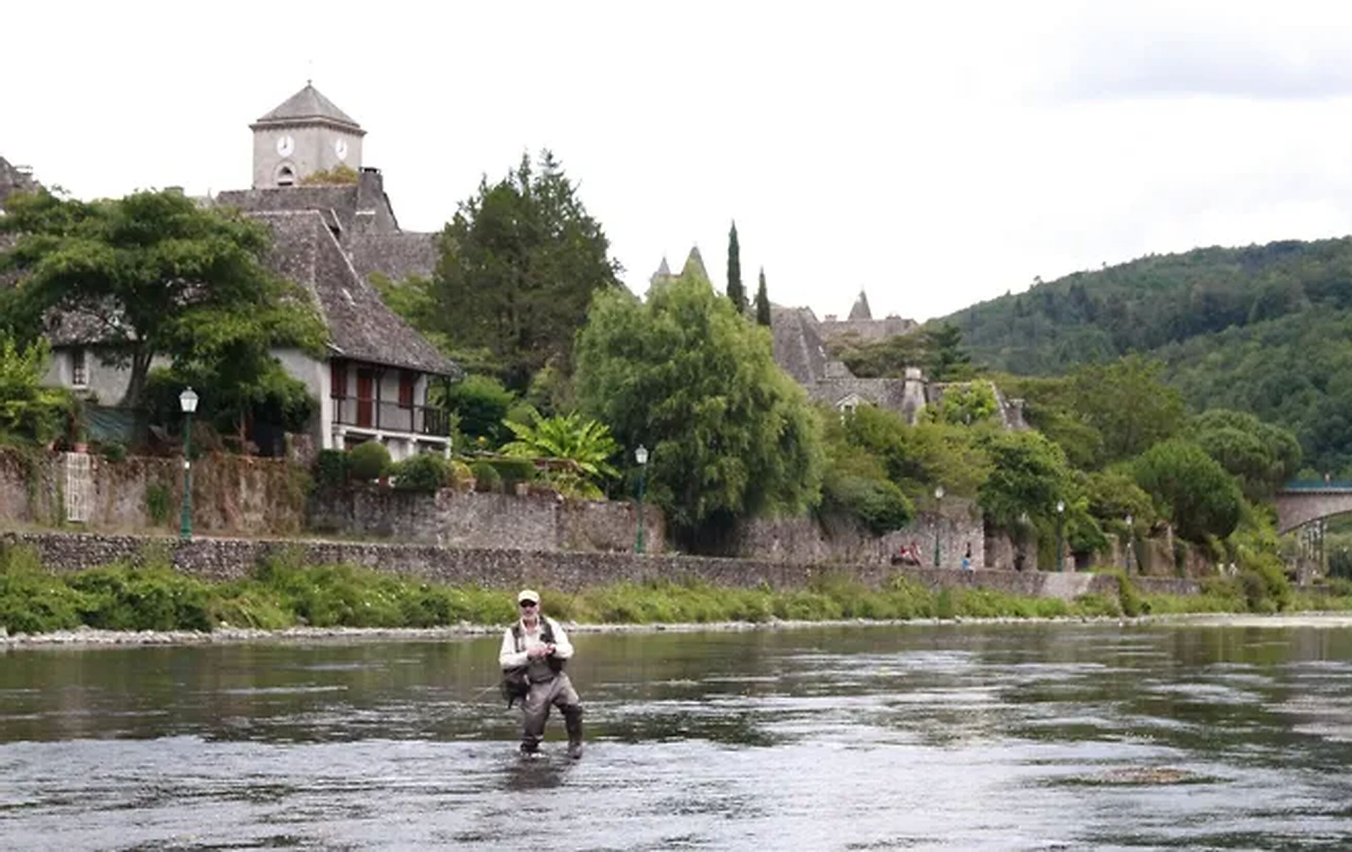 Una persona pescando en un río, con edificios y árboles al fondo.