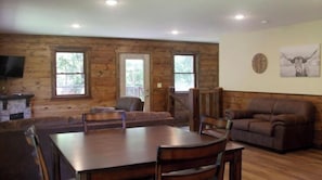 A log cabin interior with a dining table, chairs, a sofa, and a television on a stand.