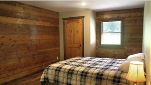 A bedroom with wooden walls, a bed with a plaid bedspread, a window with blinds, and a door.