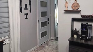 Een gang met een witte deur, een muur waarop 'EAT' in zwarte letters staat, een koffieapparaat op een plank, en een kleine potplant.
