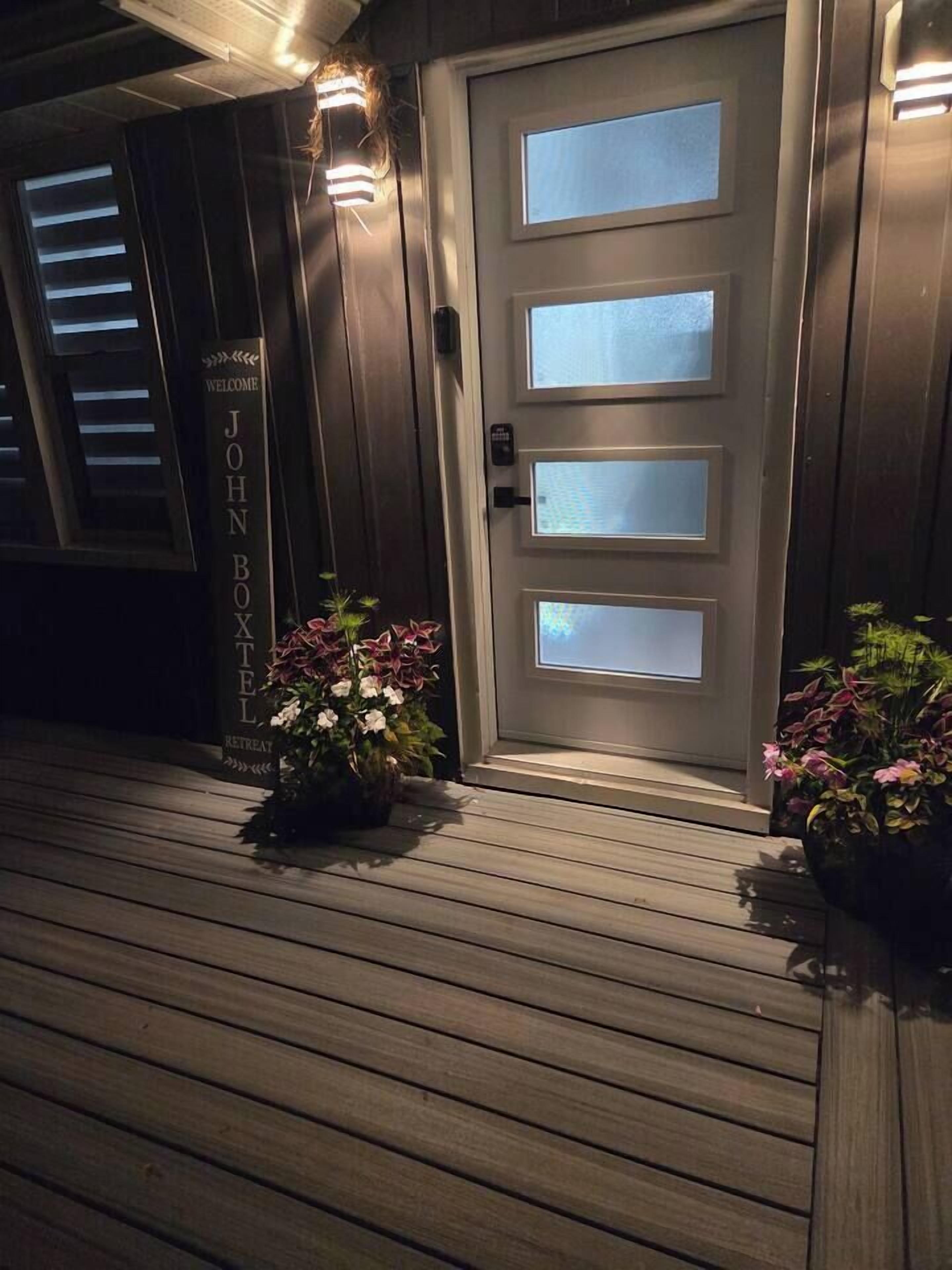 A wooden deck leading to a door with frosted glass panels, a welcome sign, and potted plants.