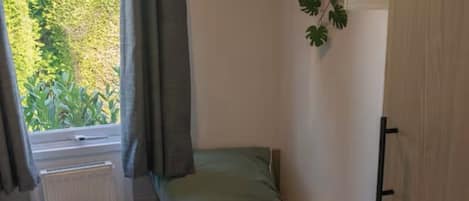 A small, compact room with two single beds, a window with curtains, a radiator, and a small plant on the wall.