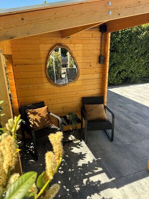 Een houten terras met een zithoek, twee stoelen, een kleine tafel, een ronde spiegel aan de muur en een haag op de achtergrond.