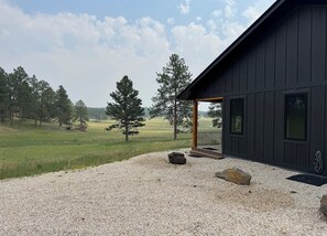 A modern building with a dark exterior, a gravel driveway, and a grassy field with trees in the background. - New build cabin with AC on 20+ acres outside Custer (Custer)