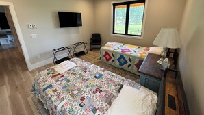 Quarto com uma cama grande, uma cama menor, uma escrivaninha com abajur, uma cadeira, uma televisão e uma janela com vista.