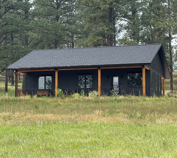 Una cabana amb teulada fosca i estructura de fusta, envoltada d'un prat herbós i arbres.