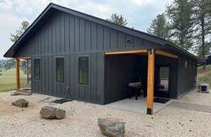 A modern building with a dark exterior, a covered porch, and a small outdoor seating area. - New build cabin with AC on 20+ acres outside Custer (Custer)