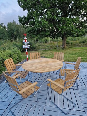 Une table ronde en bois, entourée de chaises en métal, est posée sur une terrasse en bois, elle-même bordée de verdure et d’un panneau indicateur.