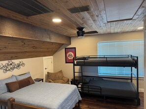 Ein Zimmer mit Etagenbett, einem Einzelbett, Holzbalken an der Decke, einem Deckenventilator, einem gerahmten Poster und einem Fenster mit Jalousien.