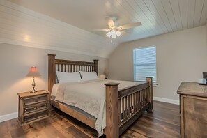 A bedroom with a wooden bed, a nightstand with a lamp, a ceiling fan, and a window with blinds. - Game day convenience and lakeside fun with is cute waterfront house (Graford)