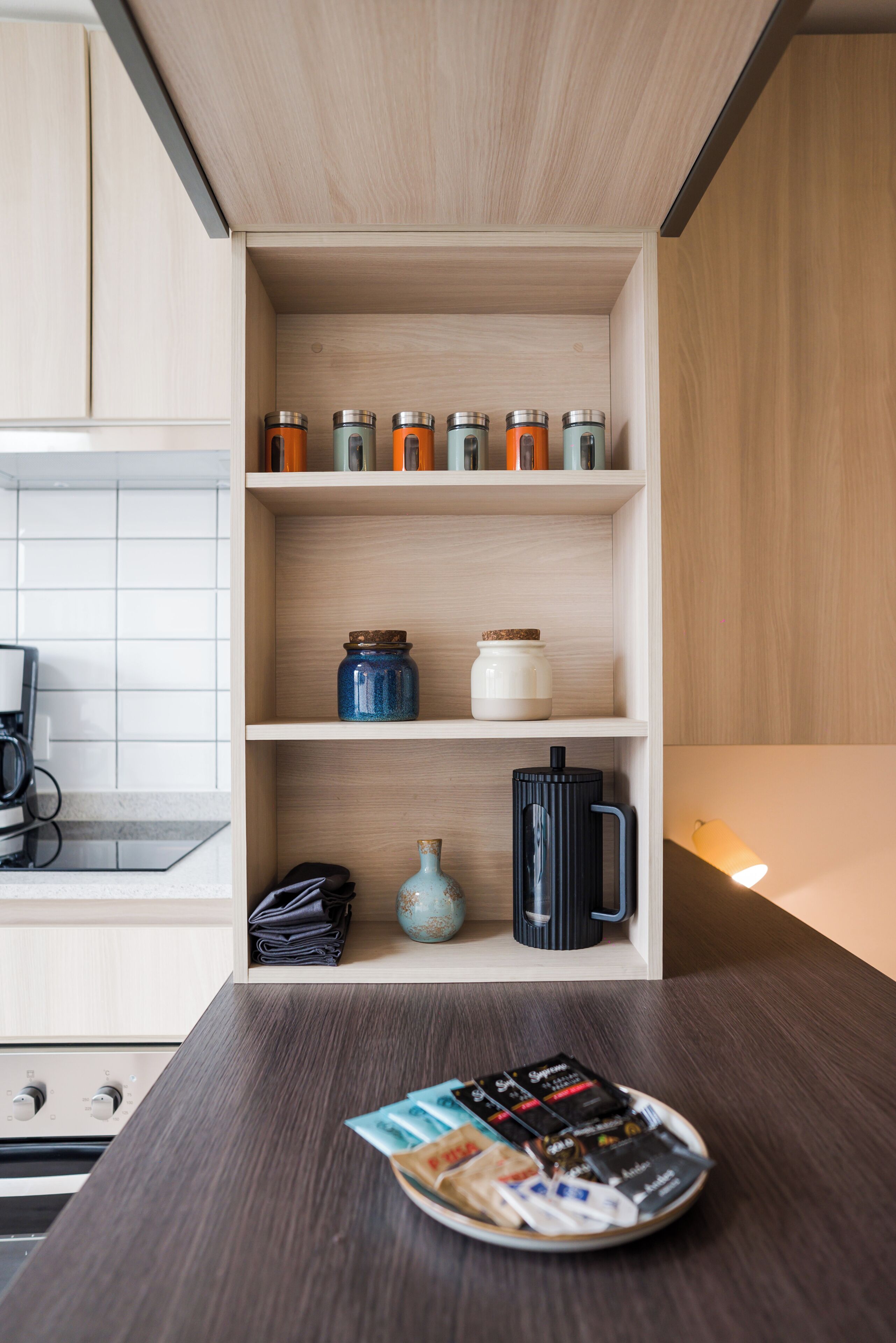 Una cocina con armarios de madera, una cafetera, una estantería con frascos y una revista sobre la encimera.