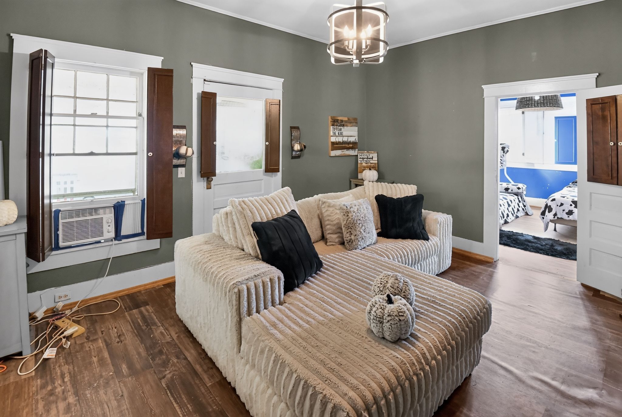 A bedroom with a large bed, a window with blinds, a chandelier, and a door leading to another room.
