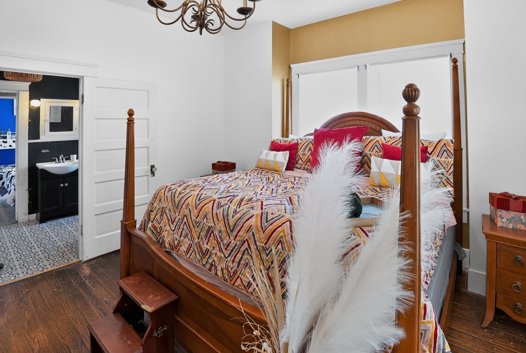 A four-poster bed with a patterned bedspread, a wooden nightstand, a bedside table, and a white feathered decorative piece.