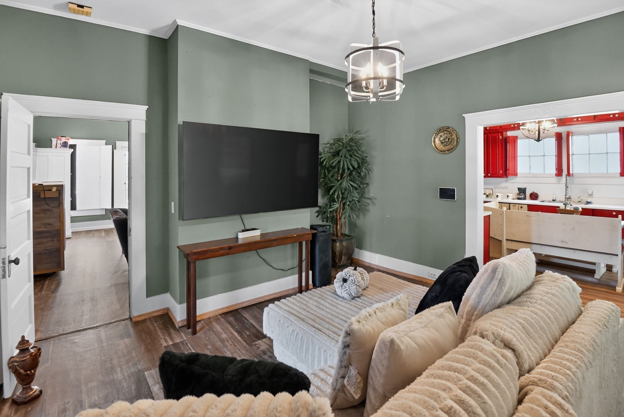 A living room with a flat-screen TV, a wooden coffee table, a beige sofa, and a red kitchen visible through an open doorway.