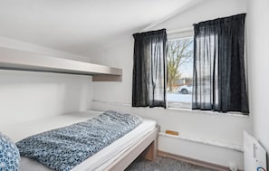 A small, single-bed room with a bunk bed above, a window with curtains, and a view of a parking area outside.