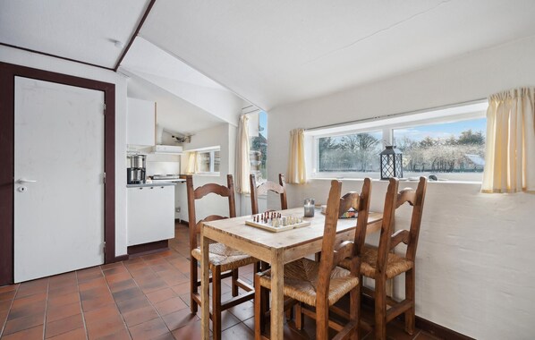 A dining area with a wooden table and chairs, a kitchen in the background, and a window with curtains.