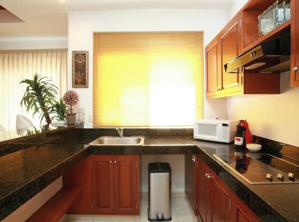 A modern kitchen with granite countertops, wooden cabinets, a stainless steel trash bin, a microwave, and a red kettle. - Cozy condo in Playa del Carmen! Unique with a Beautiful Landscape! (Playa del Carmen)