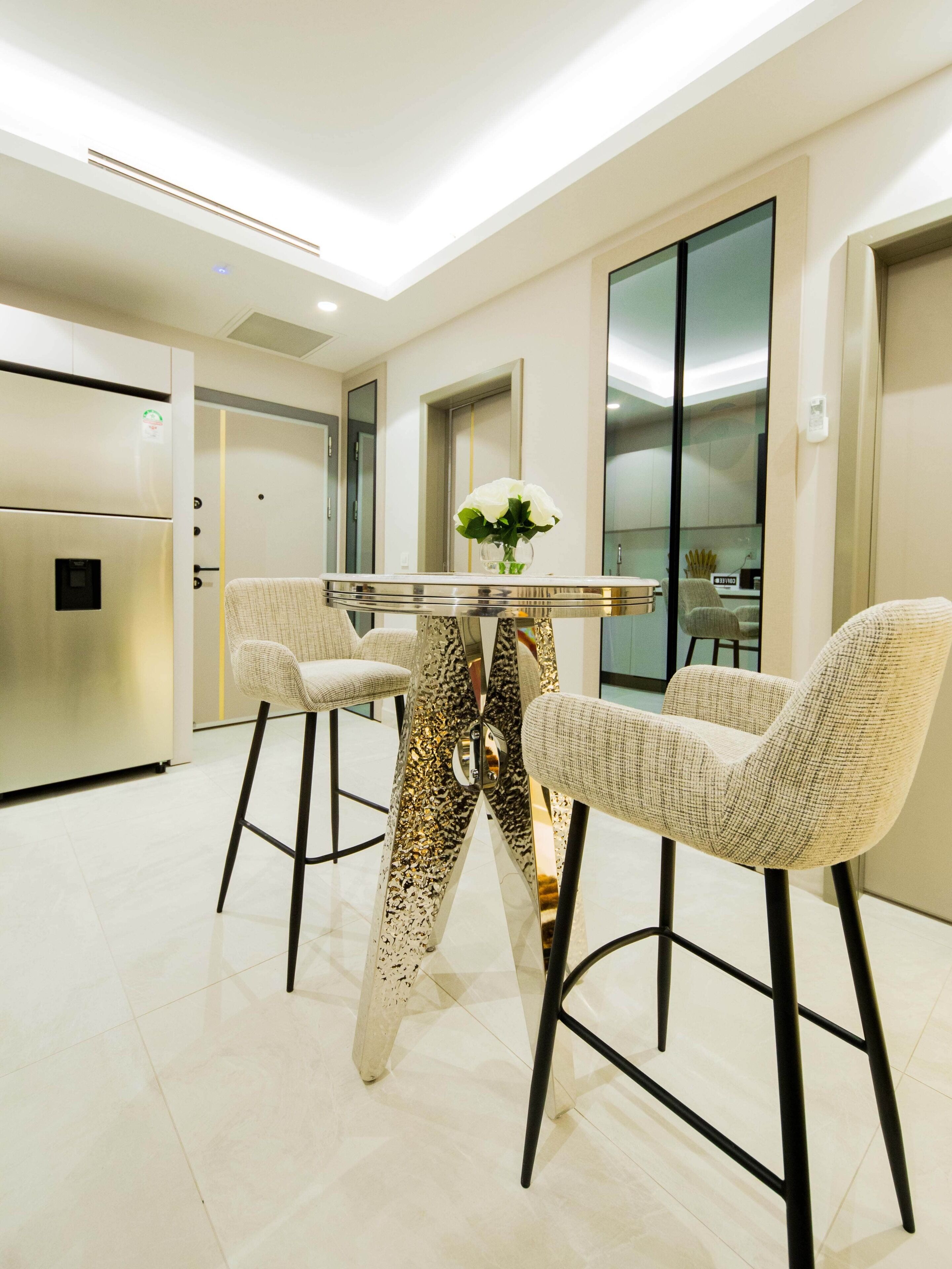 A modern kitchen with a stainless steel refrigerator, a round table with a vase of flowers, and two bar stools with a woven seat.
