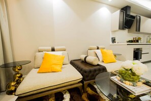 Quarto de hotel moderno com duas camas, uma mesinha, uma mesa de centro de vidro e vista para a cozinha.