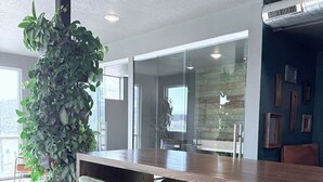 Ett modernt kontorsutrymme med ett långt träbord, två gröna barstolar, en stor krukväxt och en glasvägg.