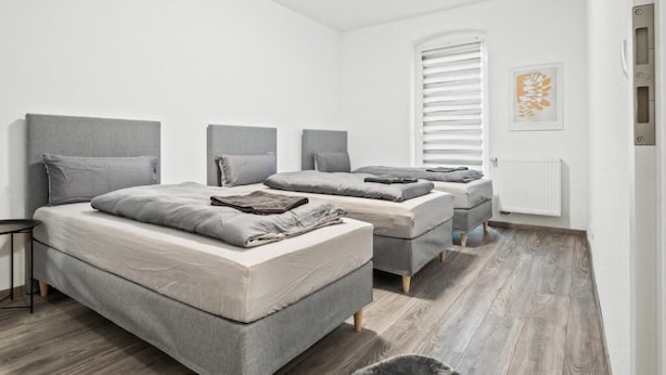 A modern bedroom with a large bed, two smaller beds, and a window with blinds. - Apartmenthaus VINA -  Smart Urban Stay (Kassel)