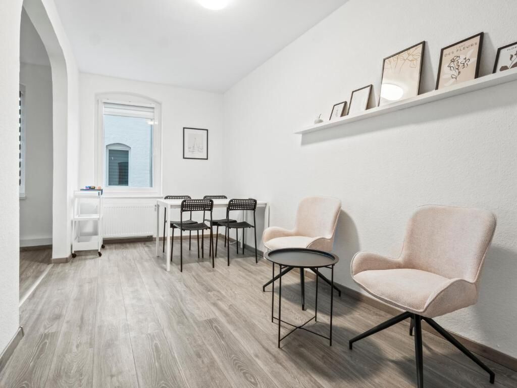 A room with a white interior, a dining area with chairs and a table, and a shelf with framed pictures.