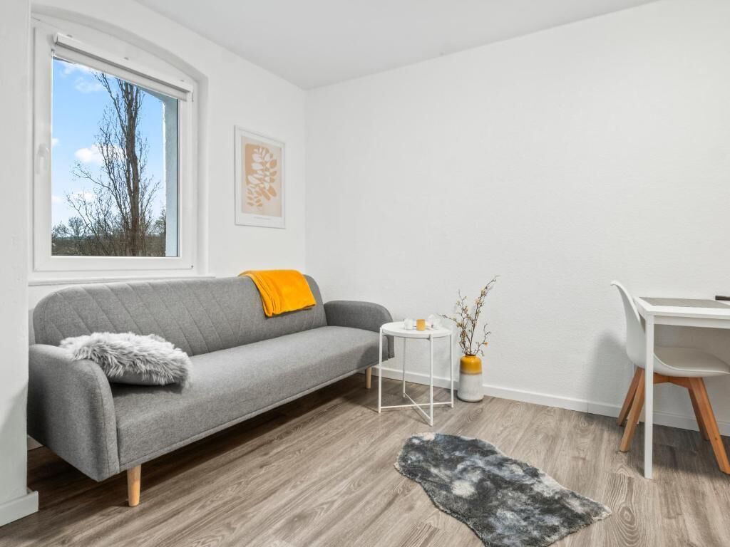 Ein modernes Wohnzimmer mit einem grauen Sofa, einem kleinen runden Tisch, einem weißen Sessel und einem Fenster mit Blick auf Bäume.