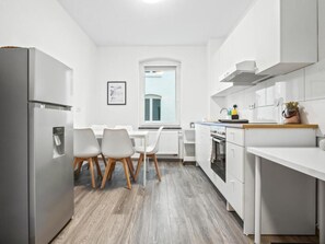 A modern kitchen with white cabinetry, a stainless steel refrigerator, a dining table with chairs, and a window with a view. - Apartmenthaus VINA - Smart Urban Stay (Kassel)