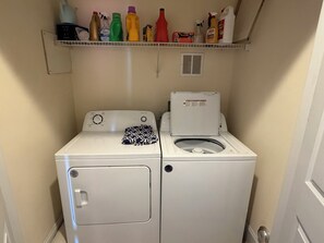 A compact laundry area with a front-loading washer and dryer, a shelf above containing various cleaning products, and a vent on the wall. - 4BR in Downtown Surf City • 1 Block to Beach (Surf City)