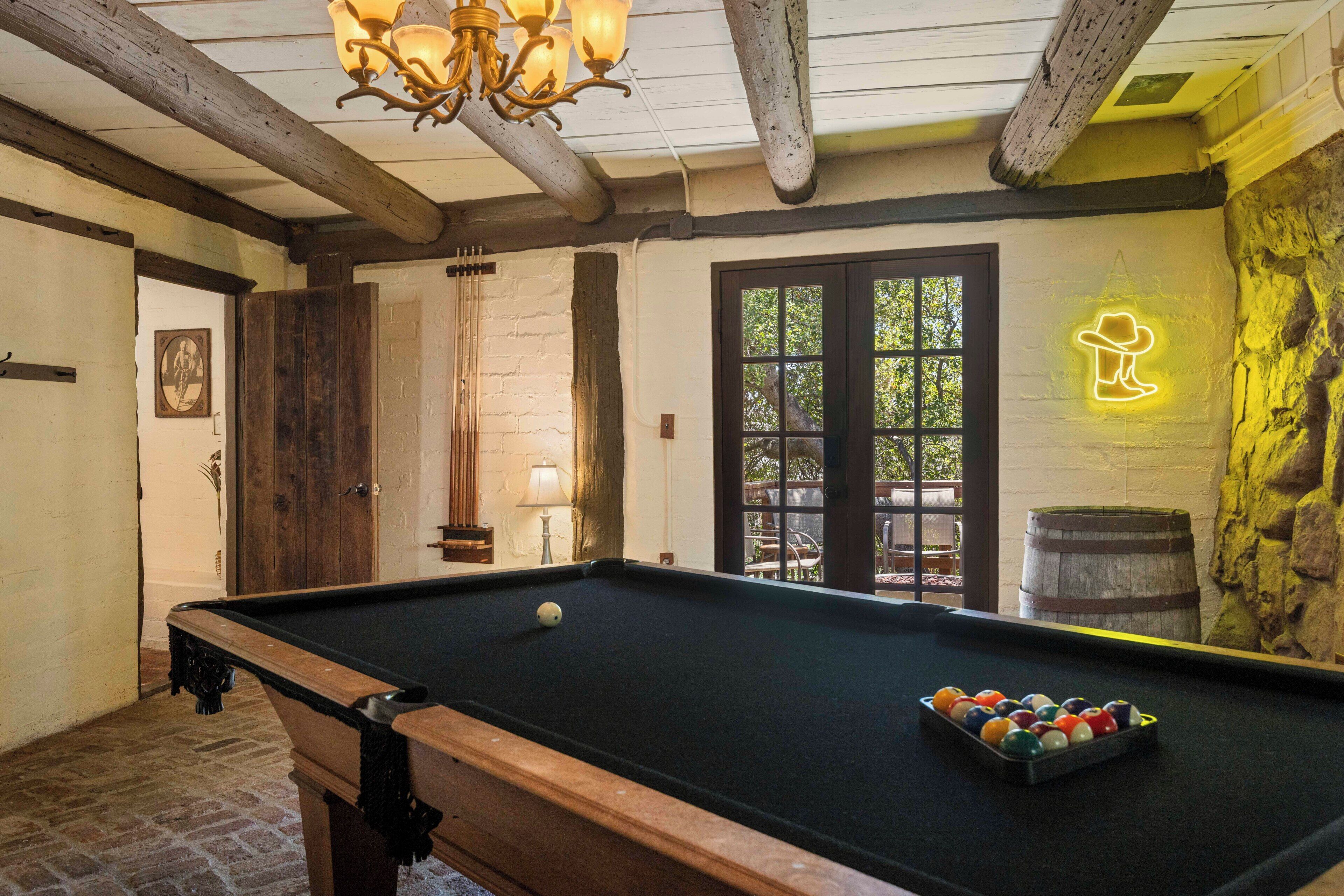 A pool table with balls, a chandelier, a wooden door, a wall-mounted neon sign, and a window with a view of the outdoors.
