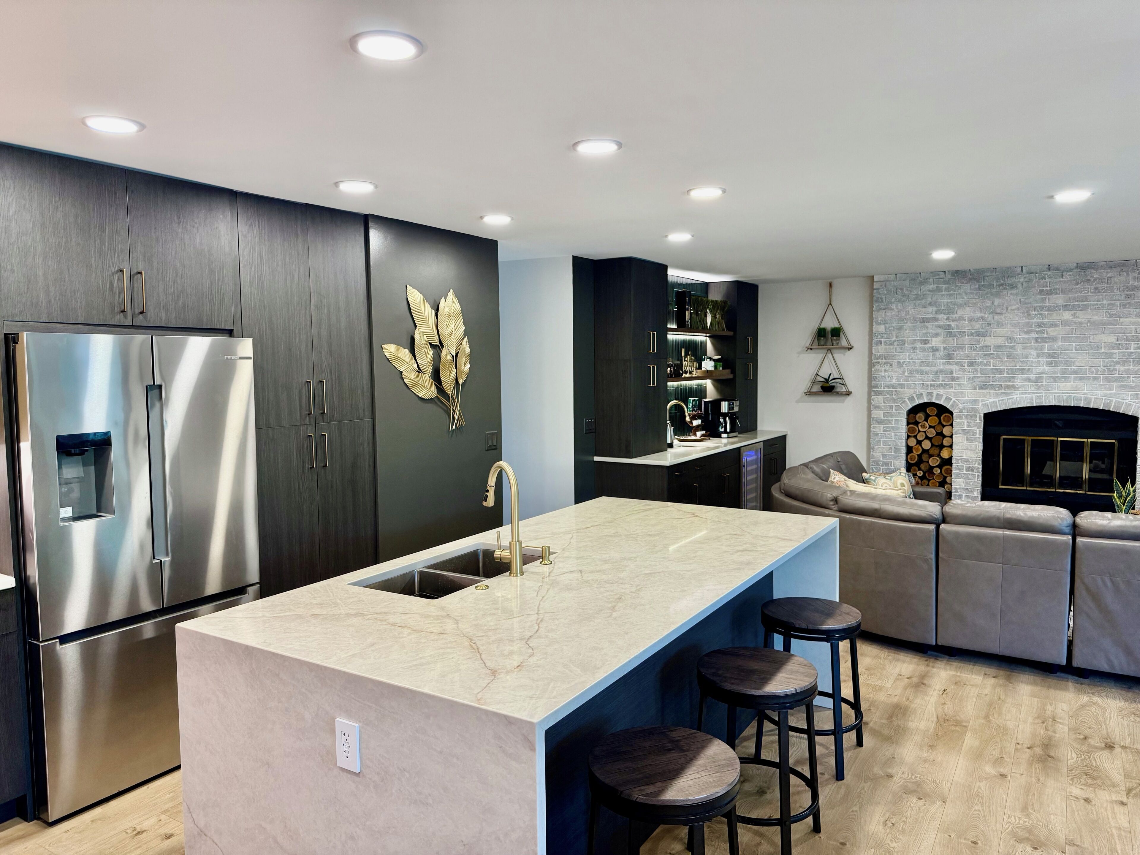 A modern kitchen with dark cabinetry, a stainless steel refrigerator, a marble island with stools, and a built-in oven.