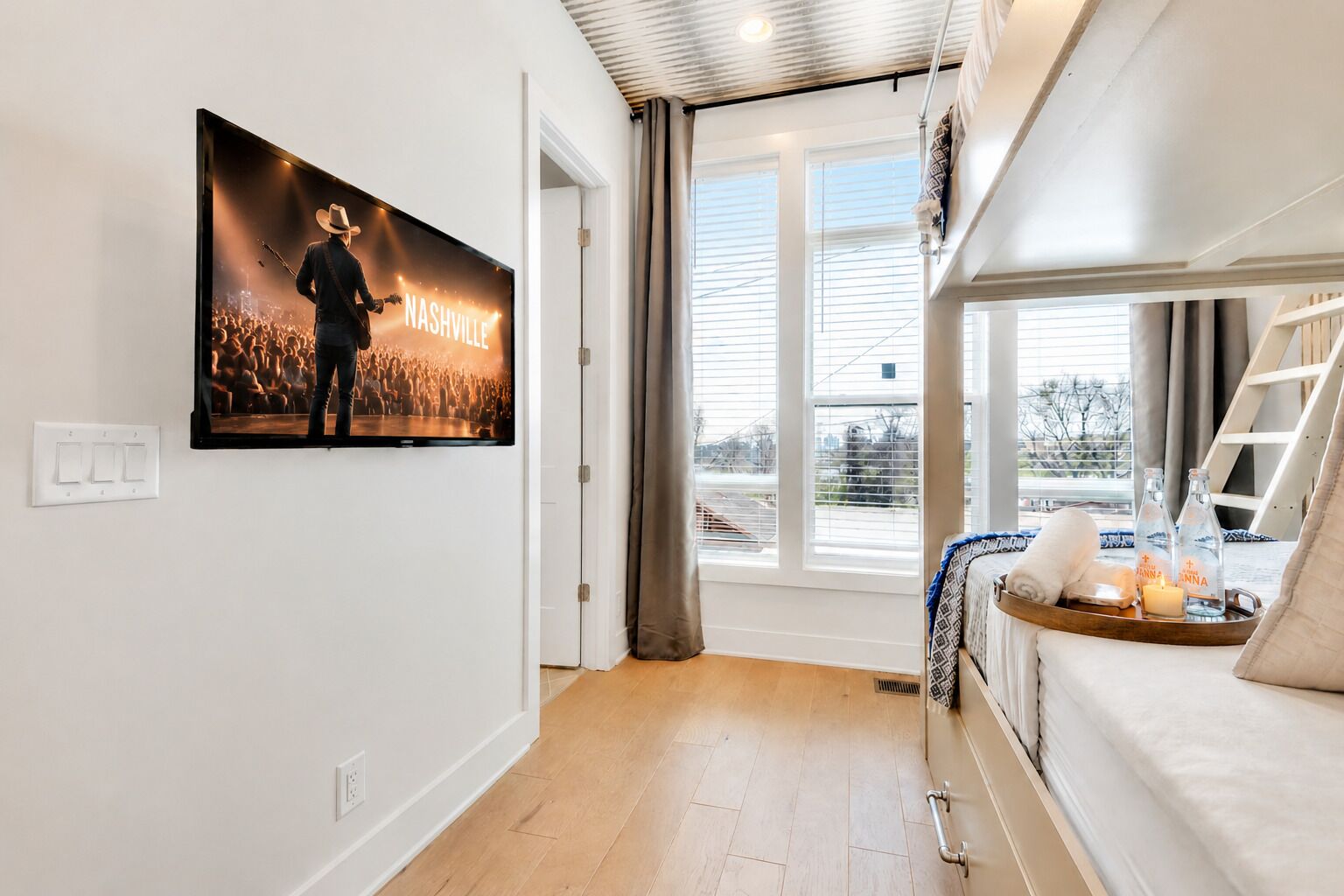 A hotel room with a large window, a bed with a headboard, a bedside table with a tray, and a wall-mounted TV displaying a concert scene.