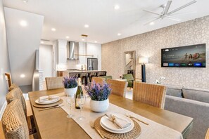 A modern dining area with a kitchen in the background, a flat-screen TV mounted on the wall, and a ceiling fan. - 2 rooftop homes near Broadway & Geodis Park, sleeps 24 (Nashville)