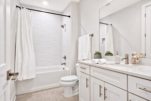 A modern bathroom with a bathtub, toilet, and vanity with a mirror and toiletries. - 2 rooftop homes near Broadway & Geodis Park, sleeps 24 (Nashville)
