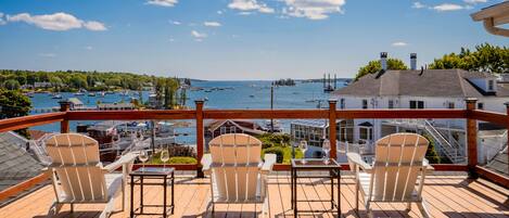 Une terrasse avec trois chaises et une table, donnant sur une marina où sont amarrés des bateaux et où se trouve un phare.