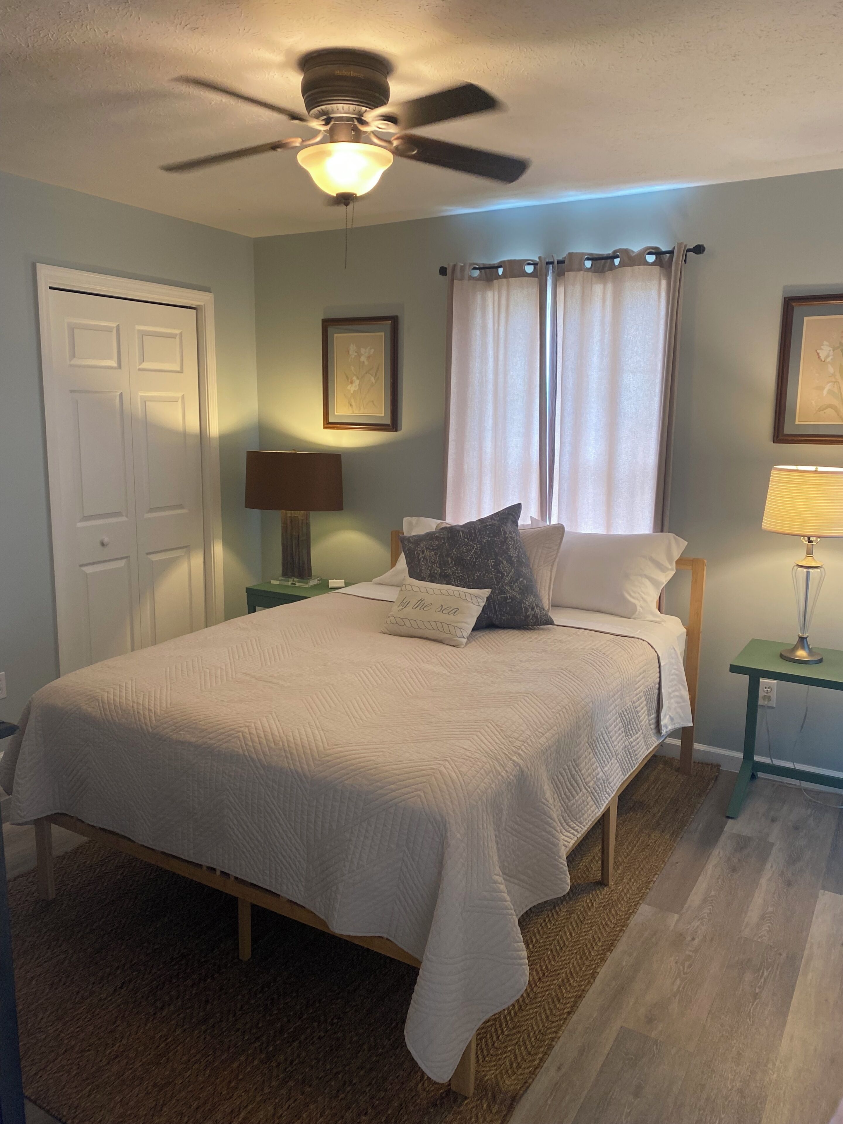 Une chambre à coucher avec un lit, un ventilateur de plafond, une table de chevet avec une lampe, une porte et une fenêtre avec des rideaux.