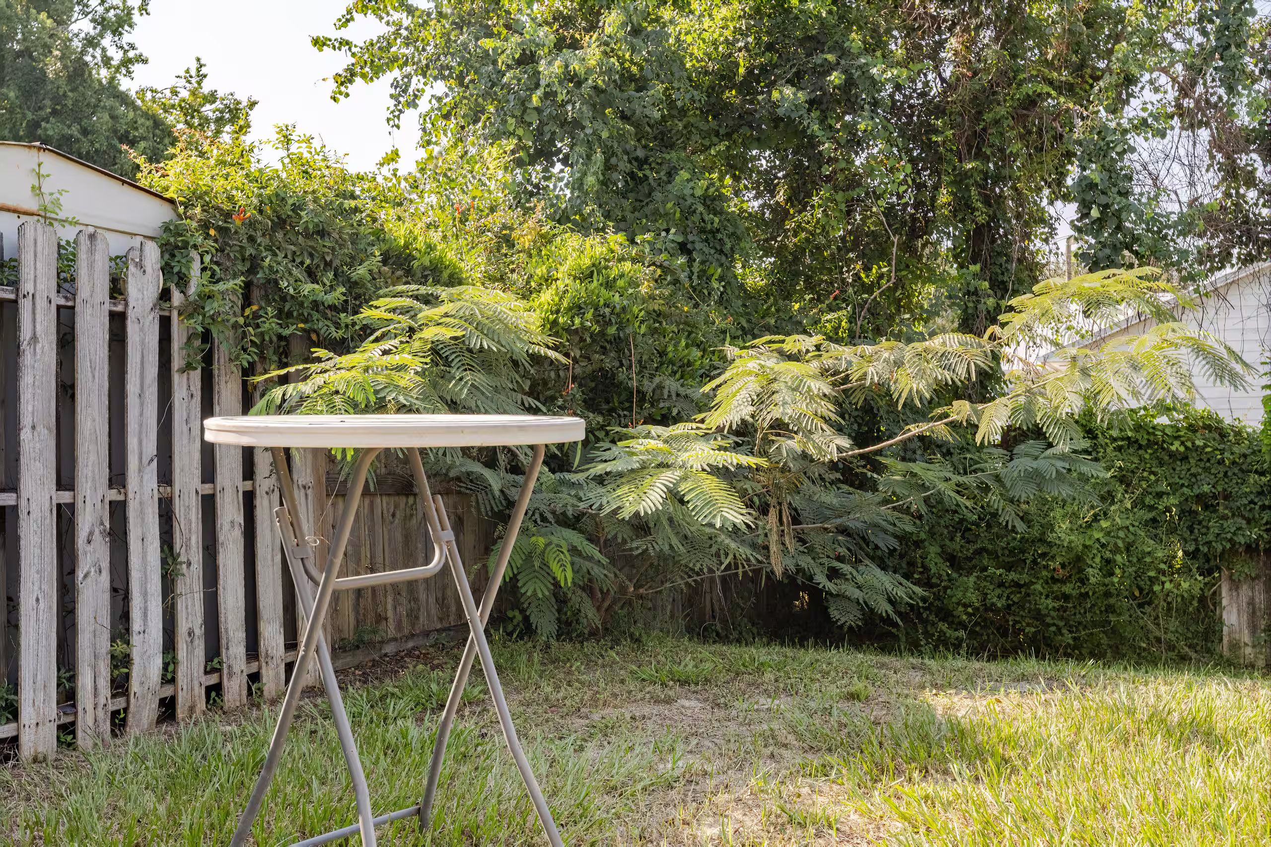 Une table ronde dotée d’un cadre en métal, placée dans une cour arrière verdoyante, avec une clôture en bois et un feuillage dense en arrière-plan.